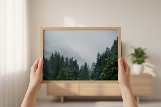 Person holding a framed picture of a forest against a neutral wall.
