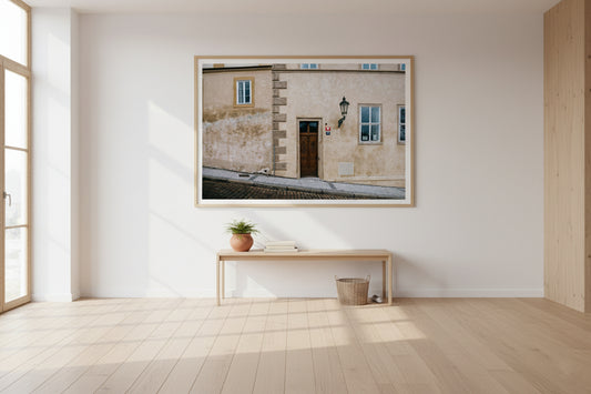Framed artwork of a building on a wall in a minimalistic room with wooden floor and large window.